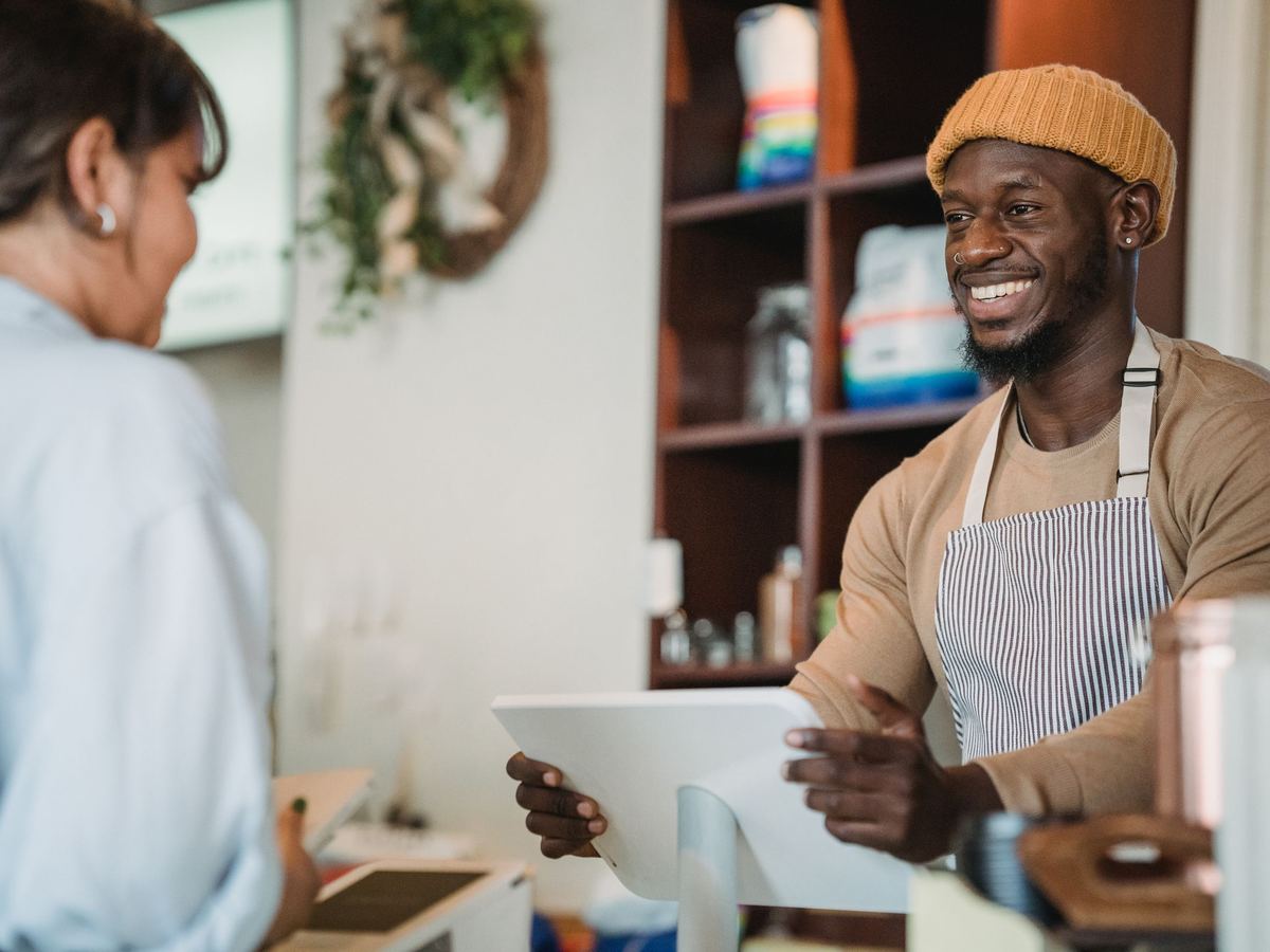 A smiling barista in an apron and beanie takes a customer's order on a tablet at a café counter.