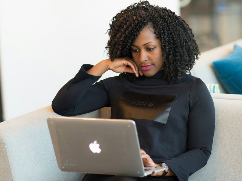 A businesswoman sits on an office sofa, working on a laptop with her chin resting on her hand.